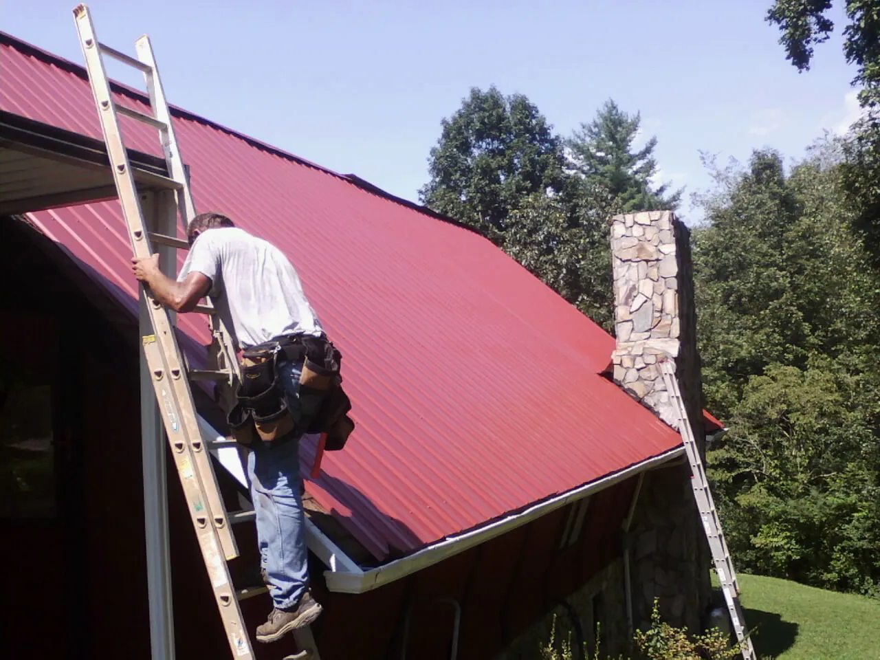 Roofer on a ladder beside a red metal roof and stone chimney — Soffit & Fascia Repair by Roofing Brothers in Princeton